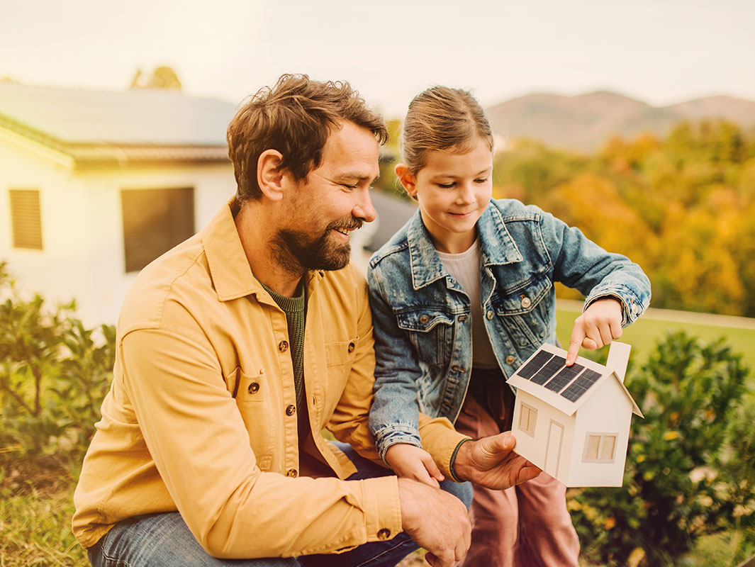 Vater und Tochter im Garten; die Tochter zeigt auf Photovoltaikanlage auf dem Dach eines Holzmodellhauses.